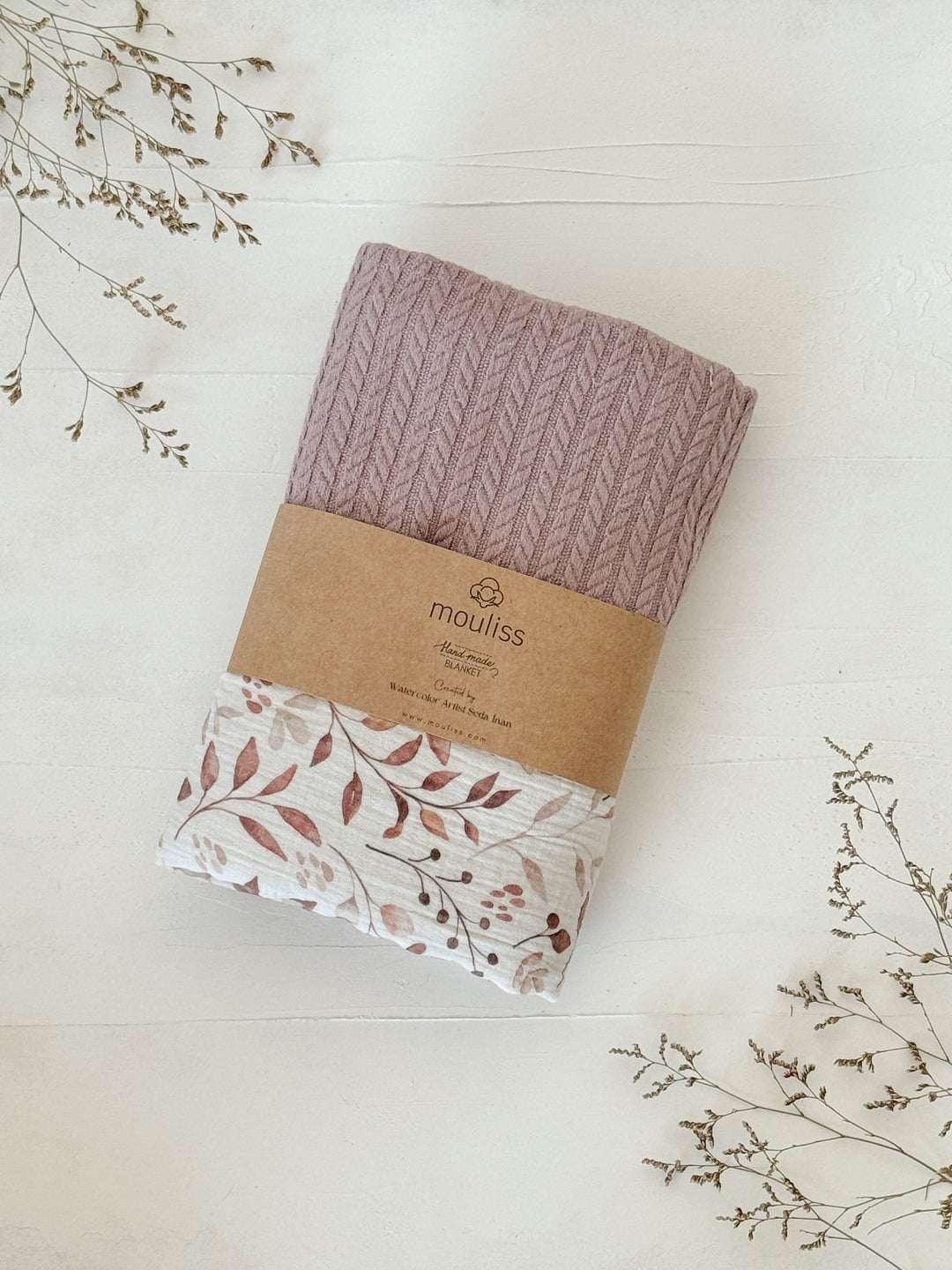 Dusty rose knitted baby blanket by Cotton, rolled to reveal a brown leaves pattern and a kraft paper band.