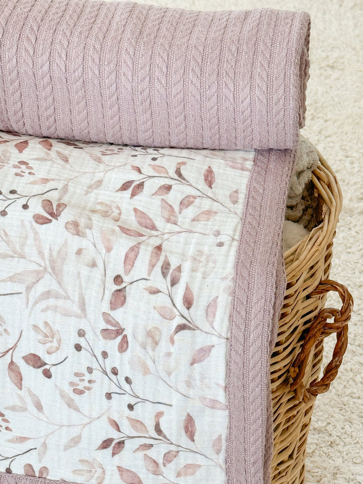 White baby blanket with brown watercolor leaves by Cotton, folded over a mauve cable-knit blanket in a wicker basket.