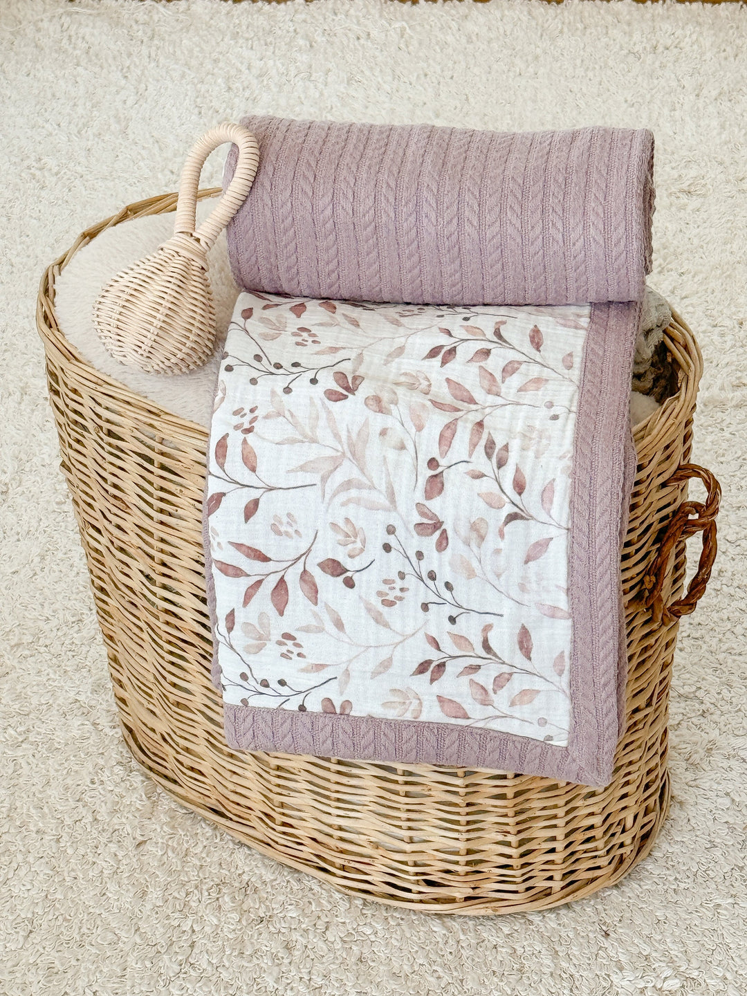 Personalized baby blanket with brownie leaves pattern by Cotton, folded with a mauve cable-knit blanket and wicker rattle in a basket.