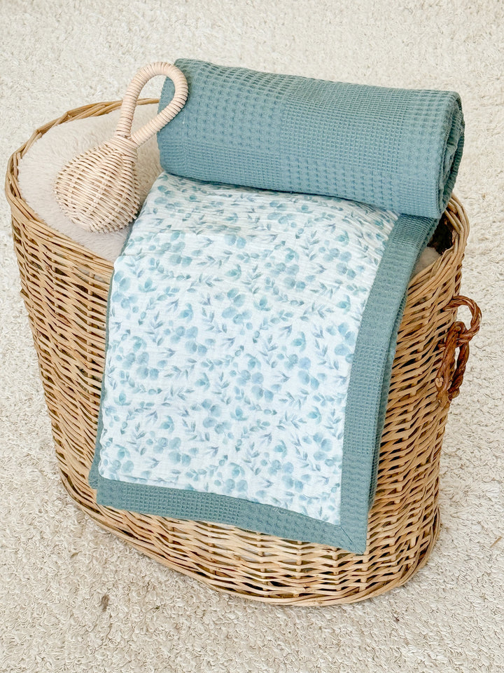White baby blanket with eucalyptus pattern by Cotton, with a blue-green waffle knit reverse, in a wicker basket with a rattle.