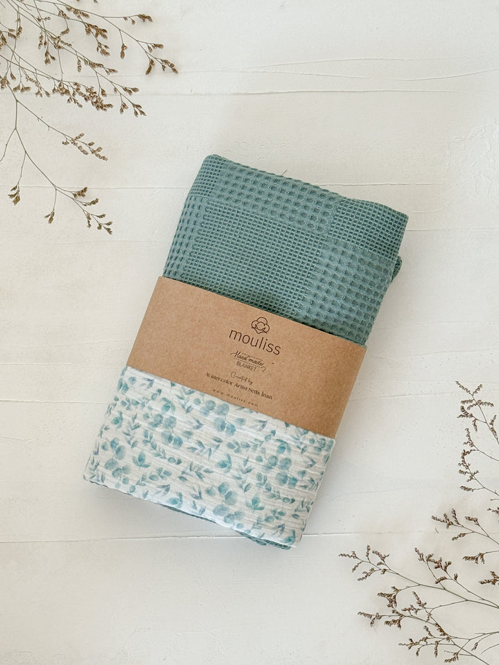Baby blanket with eucalyptus pattern by Cotton, featuring a teal waffle knit reverse, folded on a white surface with dried floral sprigs.