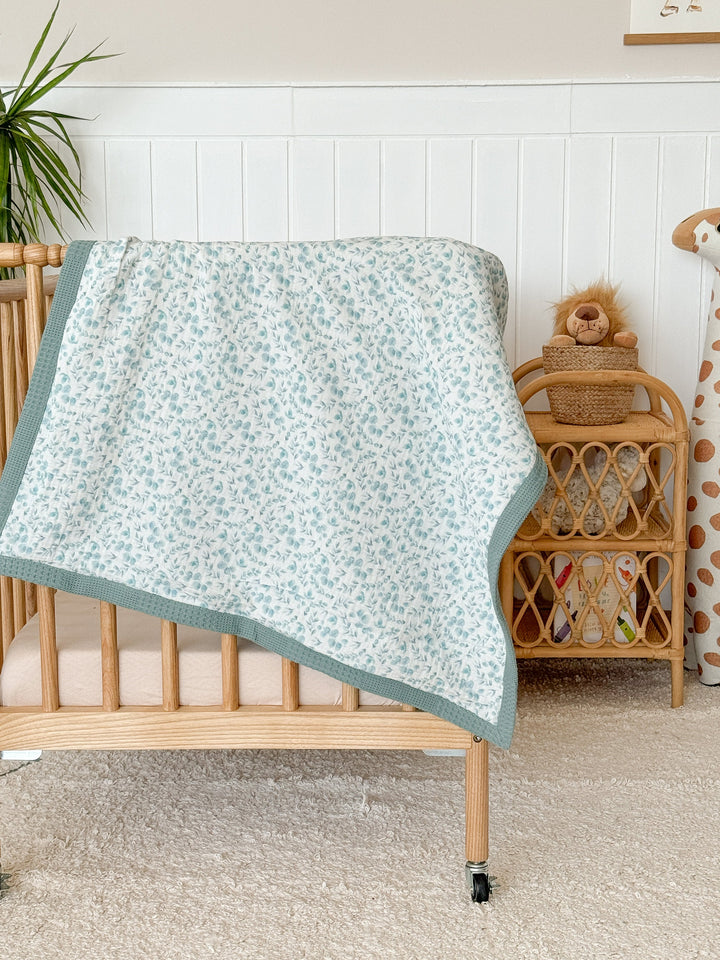 Personalized baby blanket from Cotton, featuring a light blue eucalyptus pattern on white with a teal waffle knit trim, draped over a wooden crib.