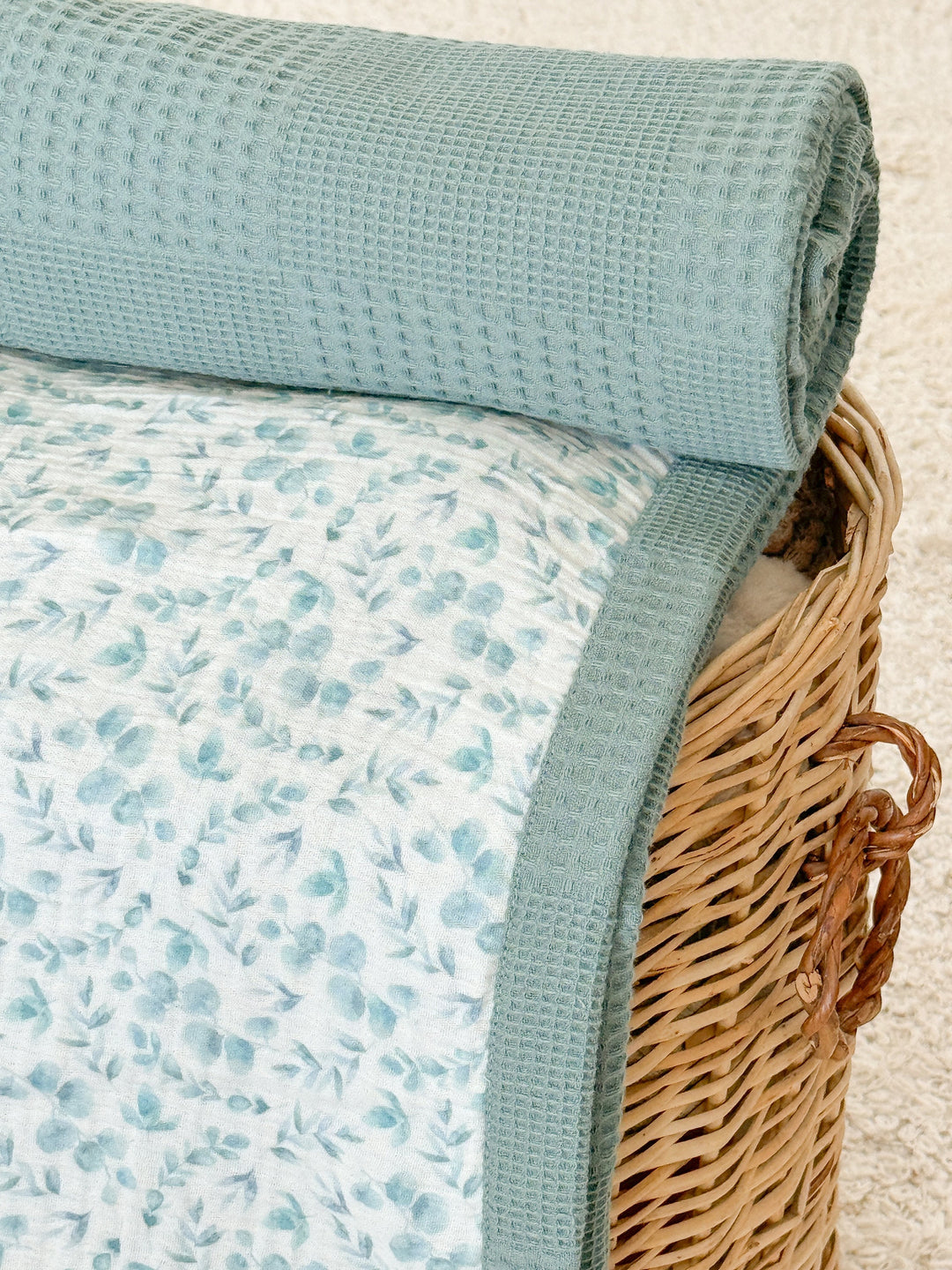 Personalized baby blanket with blue eucalyptus leaf pattern and teal waffle texture by Cotton, rolled up in a woven basket.