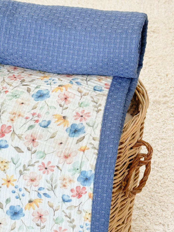 White floral and blue textured Baby Blanket by Cotton, featuring watercolor flowers and a waffle-weave texture, inside a wicker basket.