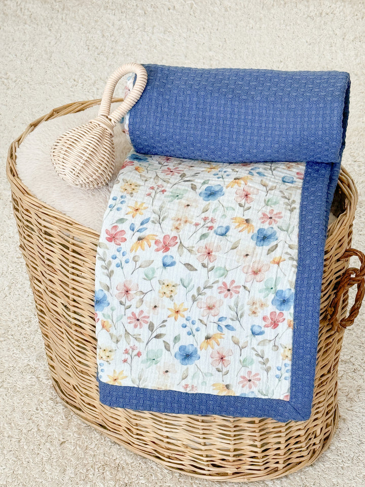 White floral and blue waffle-knit baby blanket by Cotton, folded in a woven basket with a wicker rattle on a fluffy rug.