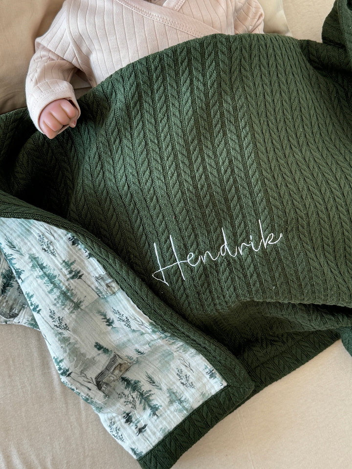 Personalized dark green cable knit baby blanket by Cotton, with a forest print lining and white embroidered name Hendrik.