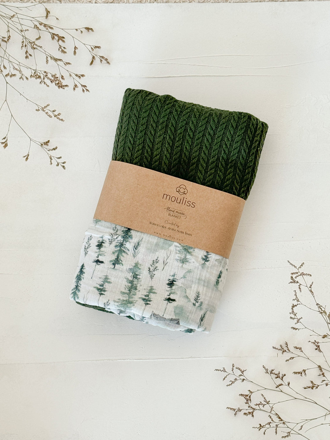 Green knitted and white patterned baby blanket by Cotton, featuring a watercolor forest pattern and a brown paper band with &