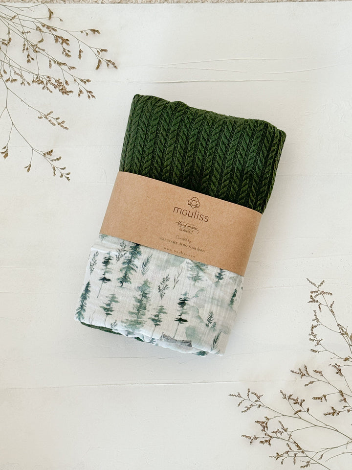 Green knitted and white patterned baby blanket by Cotton, featuring a watercolor forest pattern and a brown paper band with &