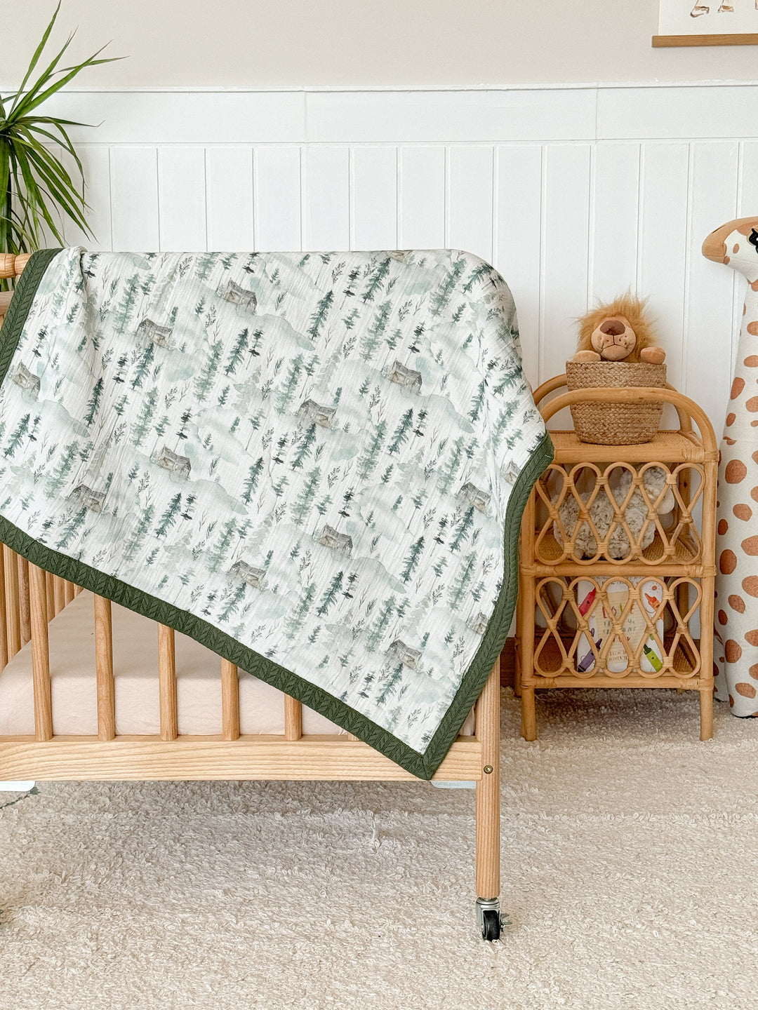 Personalized baby blanket by Cotton, featuring a pale green mountain forest pattern on crinkled muslin with a dark green border, draped on a wood crib.