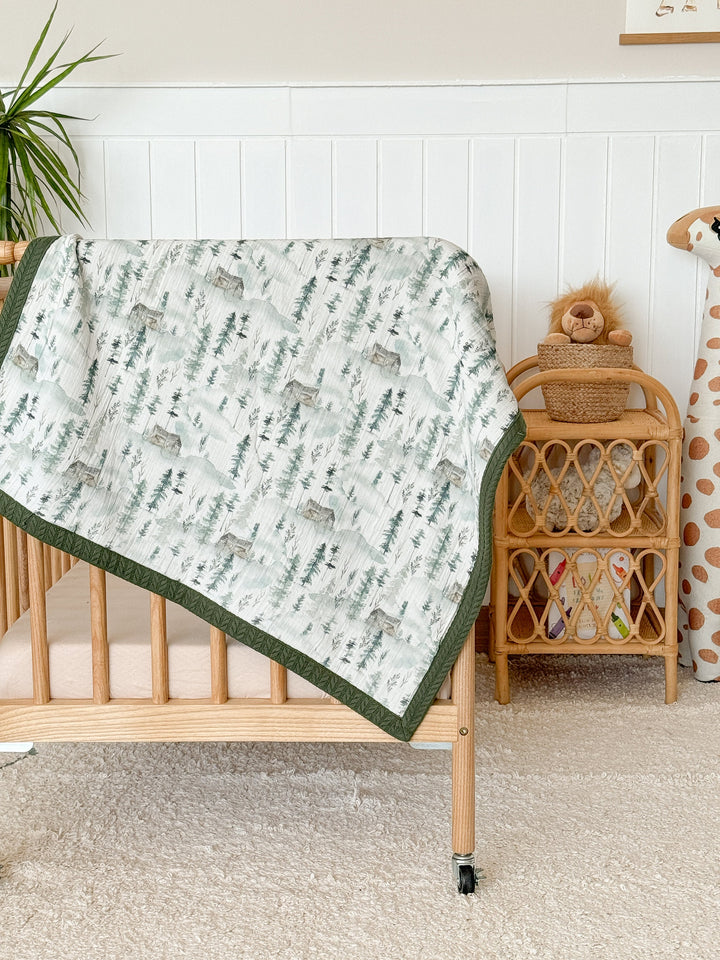 Personalized baby blanket by Cotton, featuring a pale green mountain forest pattern on crinkled muslin with a dark green border, draped on a wood crib.