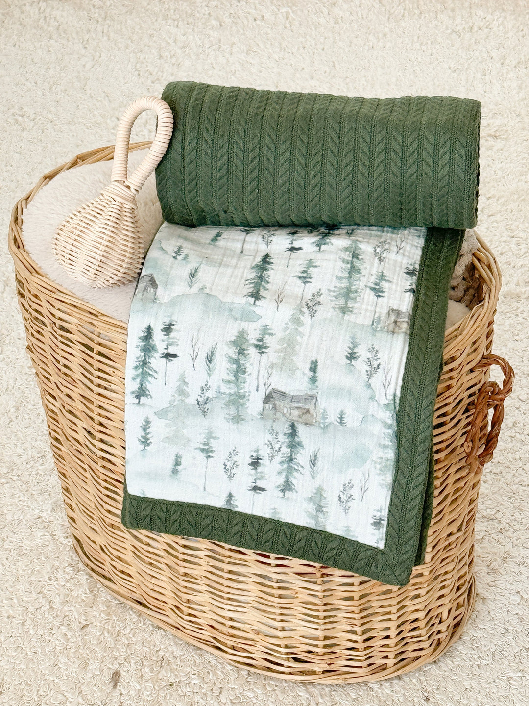 Baby blanket with High Mountains pattern by Cotton, showcasing green trees and cabins, alongside a coordinating green knit blanket and wicker rattle in a basket.