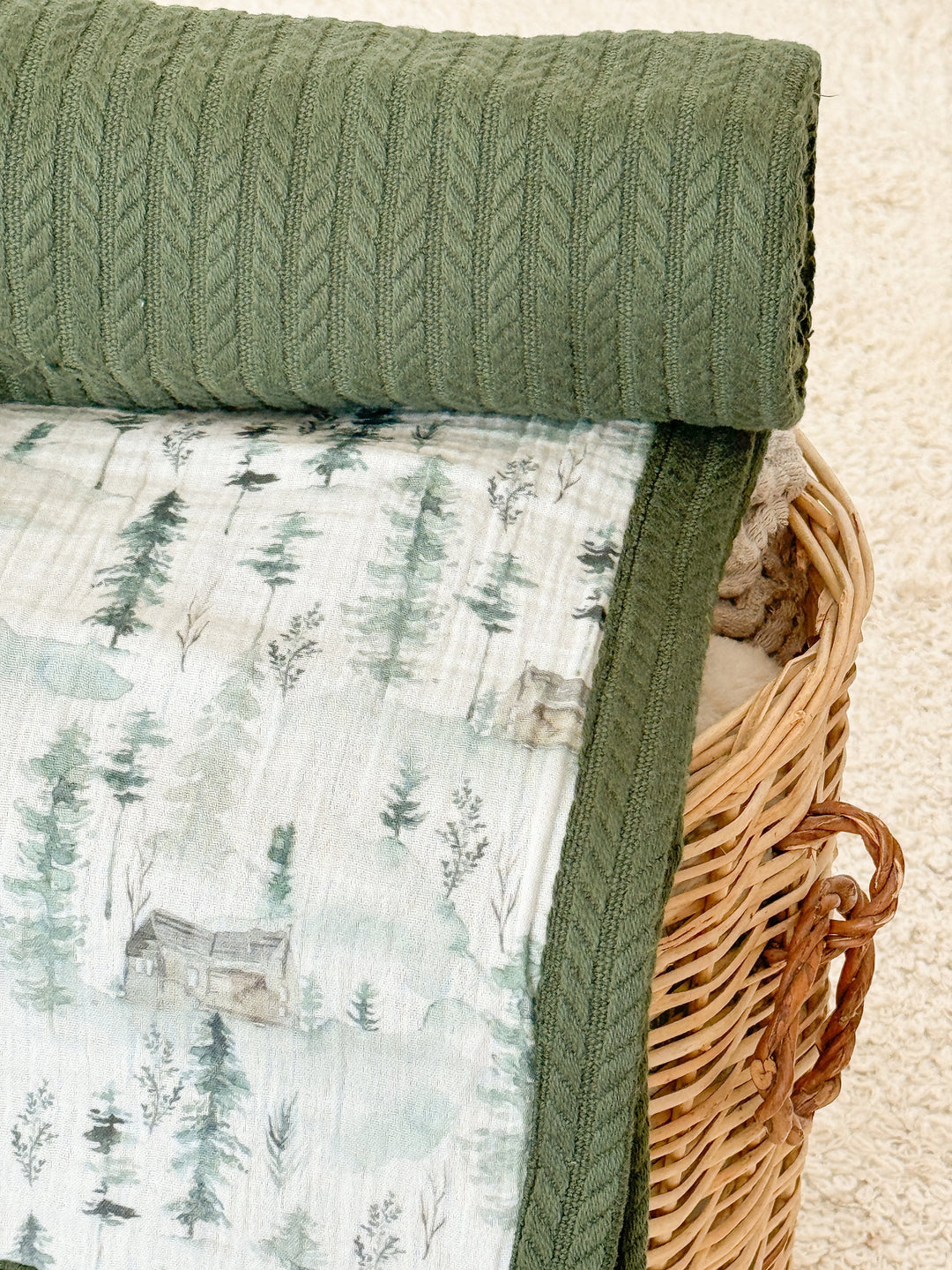 White muslin baby blanket by Cotton with watercolor forest and mountain pattern, folded under a rolled green knit blanket with herringbone texture, in a wicker basket.