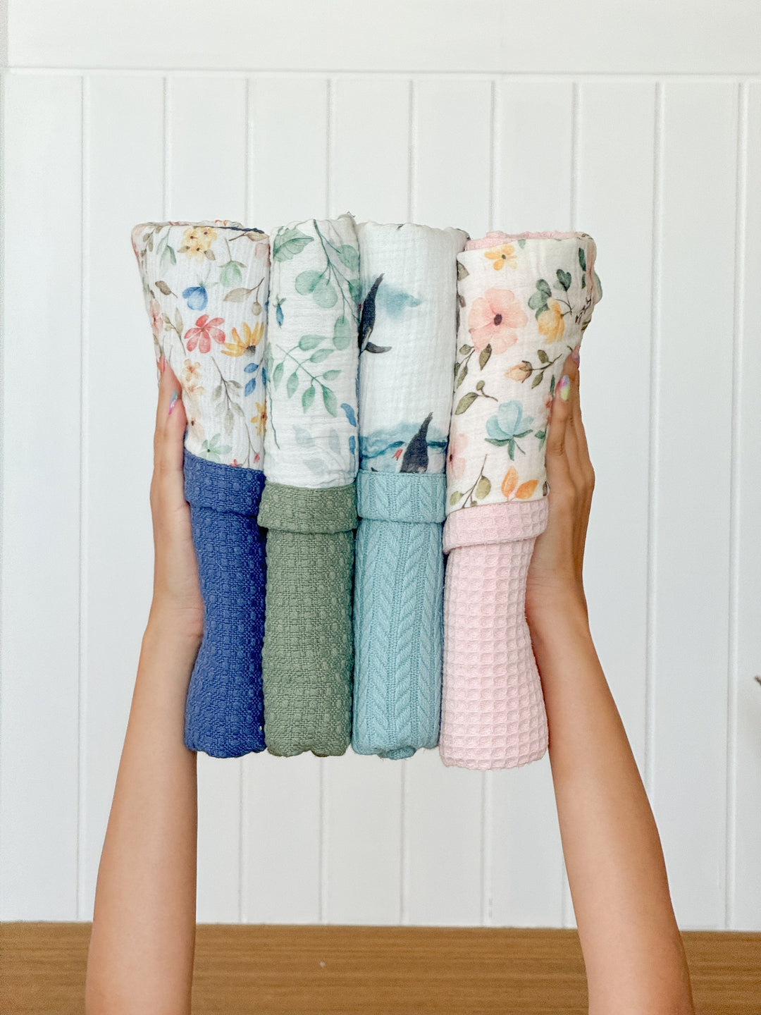 Four rolled baby blankets by Cotton, featuring diverse floral, leaf, and whale patterns with textured bottoms, held by hands.